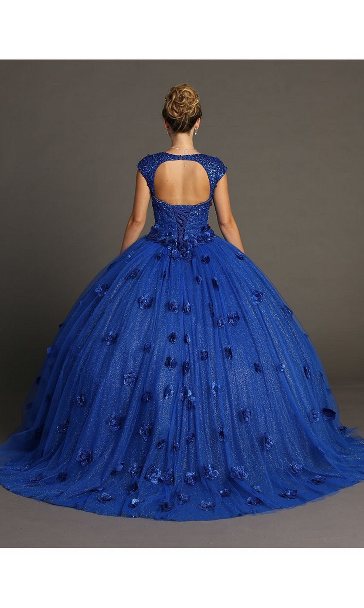 A person wearing the Quinceanera Dress CHF3068 By Chicas: a blue ball gown featuring cap sleeves, an open back, and a full tulle skirt adorned with 3D flowers and floral accents, standing against a plain background.